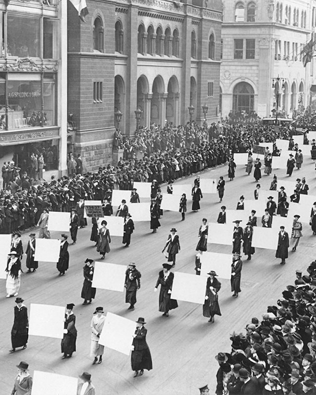 a black and white photo of US suffragists marching down a wide city street, carrying banners