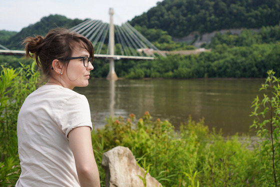 A photo of Kendra, a 30-something white woman with brunette hair, staring out across the Ohio River. There is a bridge in the background.