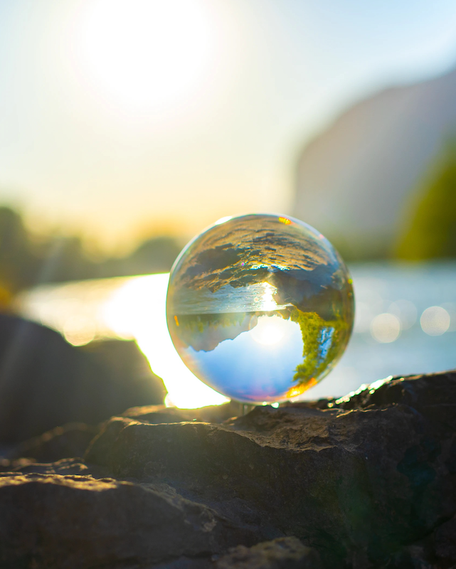 a photo of a clear marble balanced on a rock in fron of a lake, reflecting the rocks and water