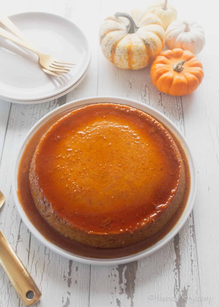 a plate of pumpkin flan on a white surface next to a plate, cutlery, and four small pumpkins