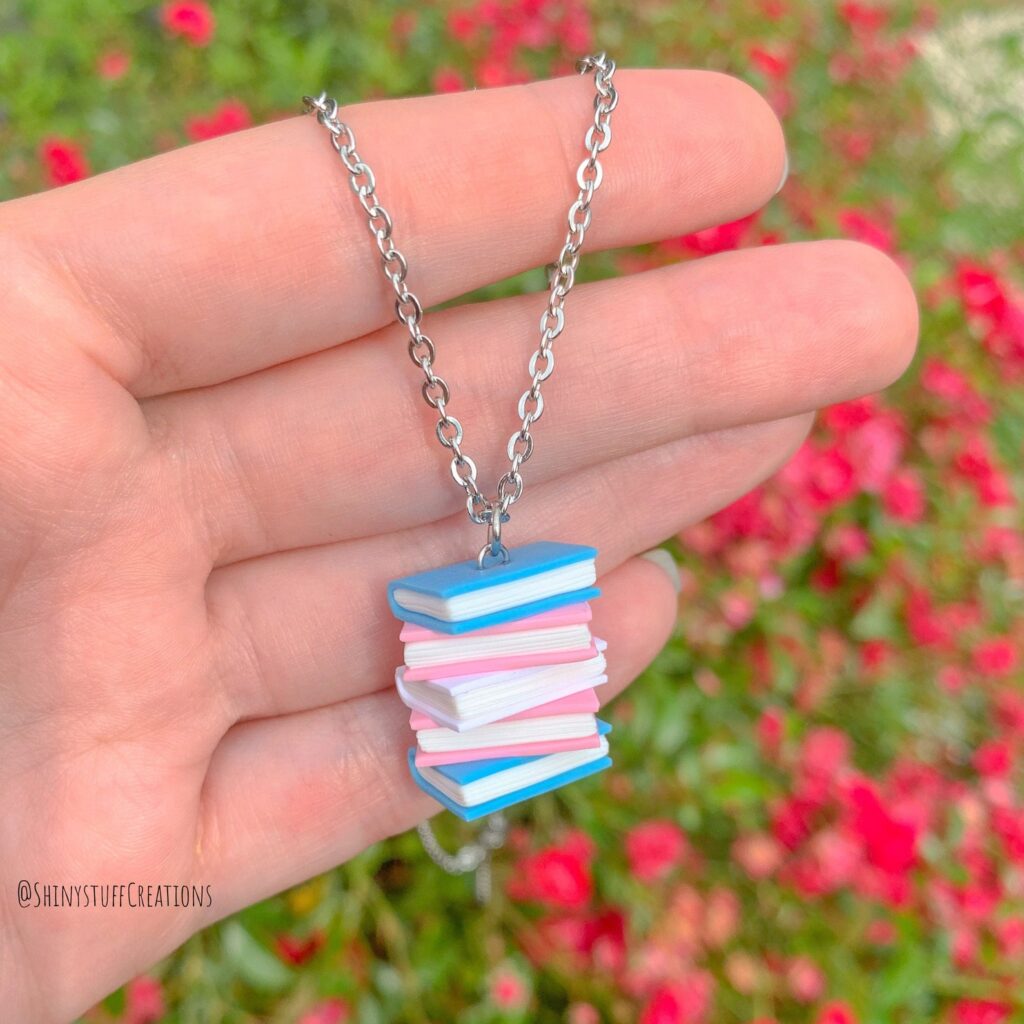 a photo of a necklace in the shape of a stack of books in the trans flag colours