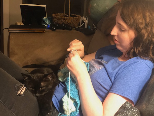 woman in a blue shirt crocheting with a black cat in her lap