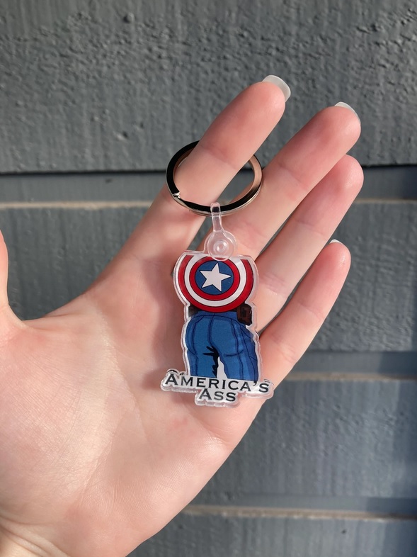 A pale hand holds on a keychain with a drawing of Captain America's butt and the text "America's Ass"
