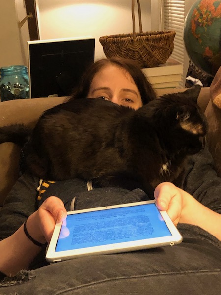 woman laying down on a couch with an ereader, and a black cat sitting on her chest and blocking her view