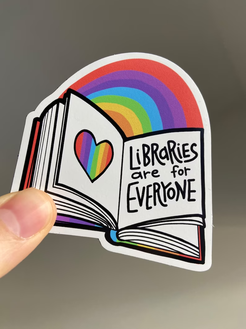 a photo of a magnet with a rainbow and an open book reading Libraries are for Everyone being held up by a hand with light skin