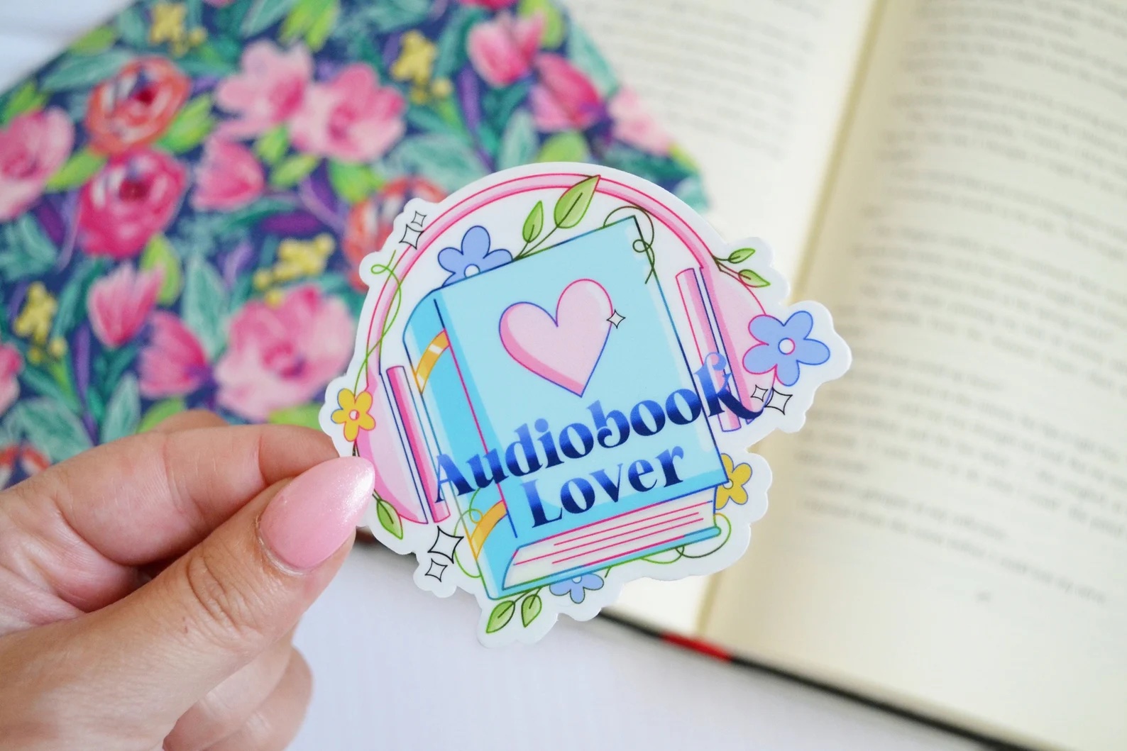 A photo of a pastel sticker featuring a book with headphones over it. The text on the sticker says, "Audiobook Lover"