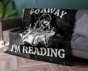 large black blanket with a white screenprinting of a skeleton reading and text "go away I'm reading"