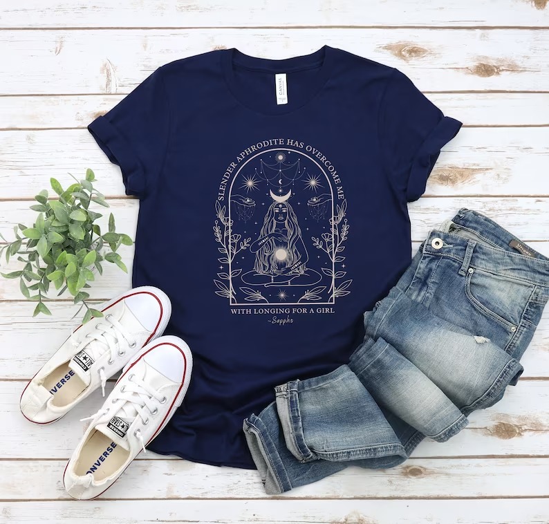 a photo of a shirt with a line art illustration of a woman, plants, and constellations with the text "Slender Aphrodite has overcome me with longing for a girl" by Sappho