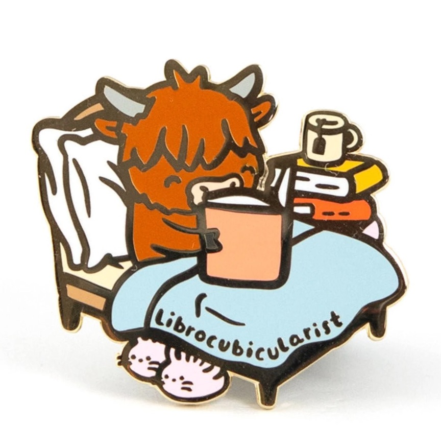 Image of an enamel pin featuring a brown cow reading in bed