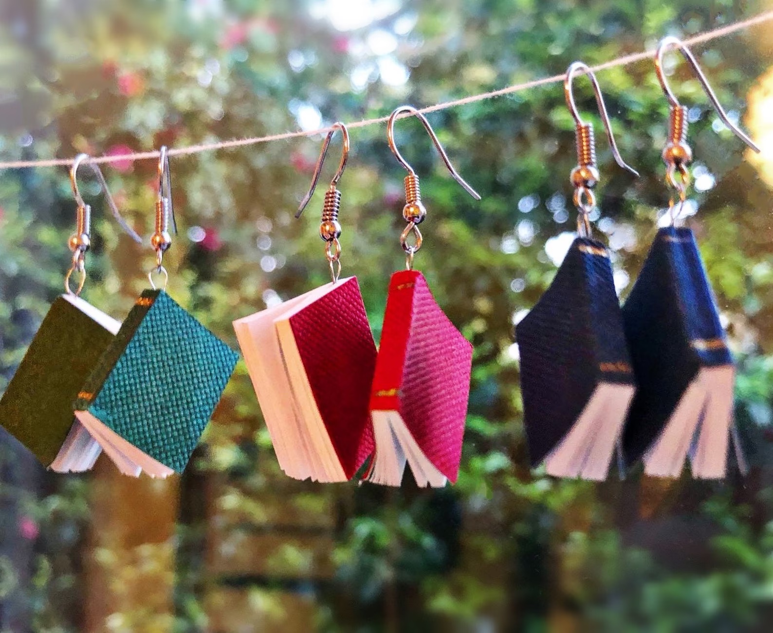 a photo of three pair of mini book earrings hanging from a strings. they are dangly earrings in red, blue, and green