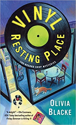 cover of Vinyl Resting Place: The Record Shop Mysteries by Olivia Blacke; illustration of a record featuring the title hanging over a shop window with a table and chairs inside, plus an orange cat and a smashed record on the sidewalk outside