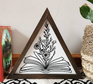 a wooden triangle frame with a black and white outline image painted inside of an opened book with flowers growing out