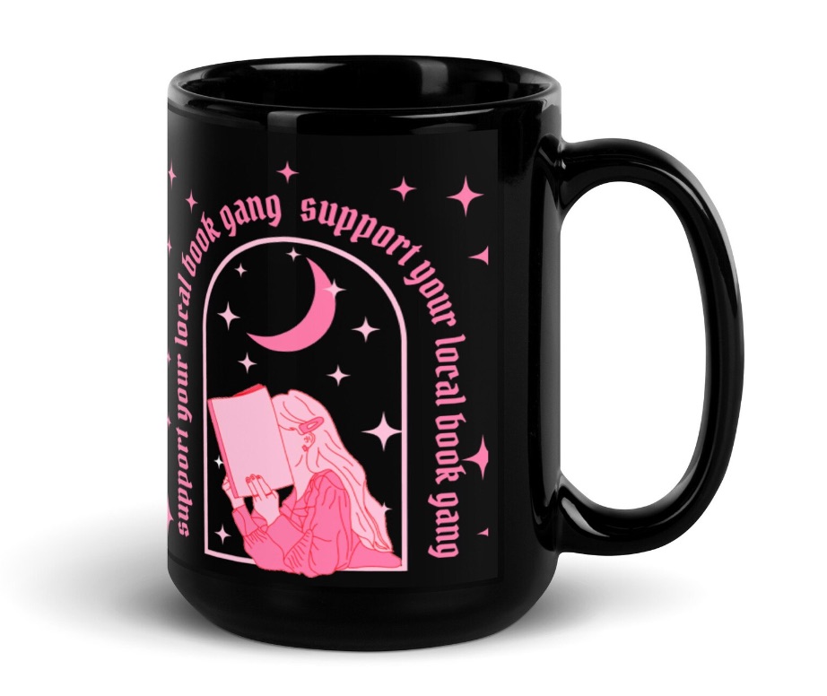 Image of a black mug with pink text that reads "support your local book gang." It has an image of a person with long hair under the text reading aa book.