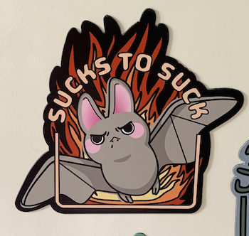 a photo of a magnet with an illustration of an adorable angry bat flying away from flames with the text Sucks to Suck