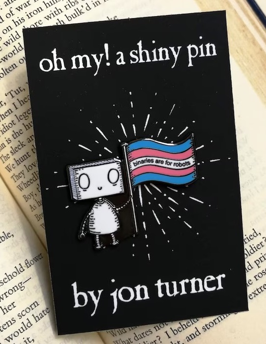 an enamel pin of a robot holding a trans pride flag with the text "binaries are for robots"