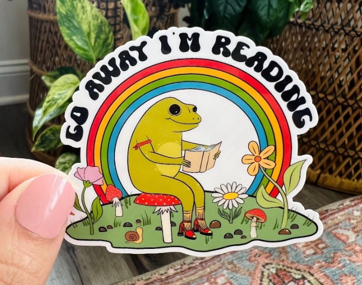 Image of a frog under a rainbow. The frog is sitting on a toadstool. Black text above the rainbow reads "Go Away, I'm Reeding."