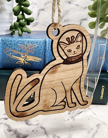 a  photo of a wooden Space cat ornament. The cat has antennae, two tails, and is wearing a helmet
