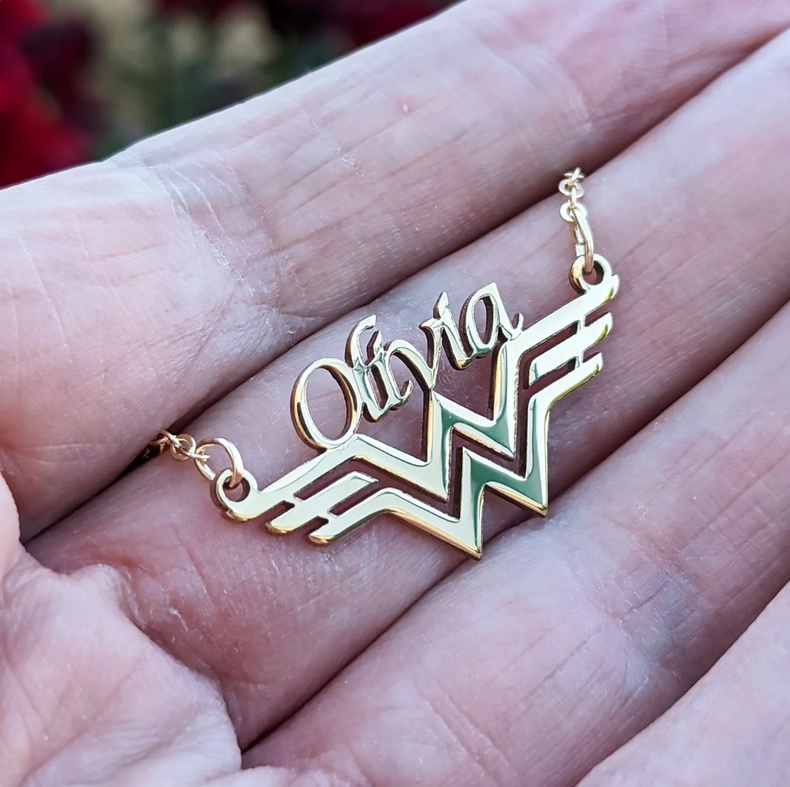 A gold pendant shaped like Wonder Woman's logo with the name "Olivia" sitting on top of it