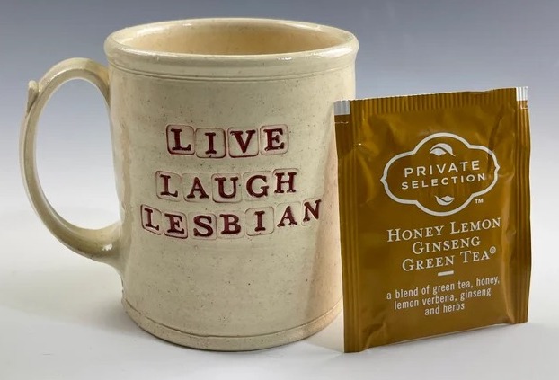 a ceramic mug stamped with letters that read Live Laugh Lesbian. A tea bag is beside it.