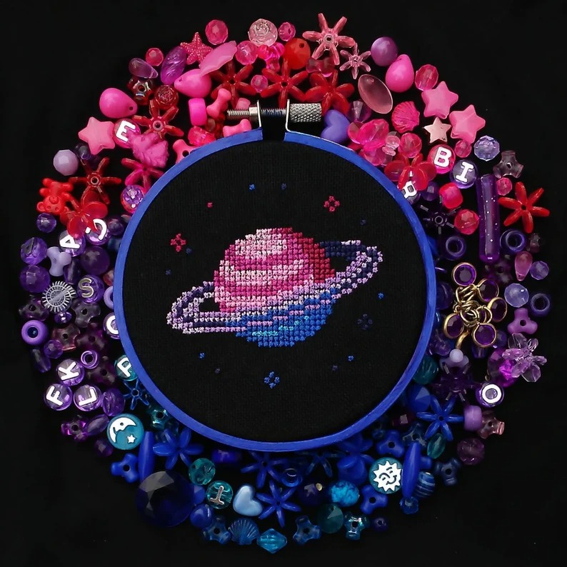 a photo of a Bi Pride Planet Cross Stitch in a hoop surrounded by matching bead colors