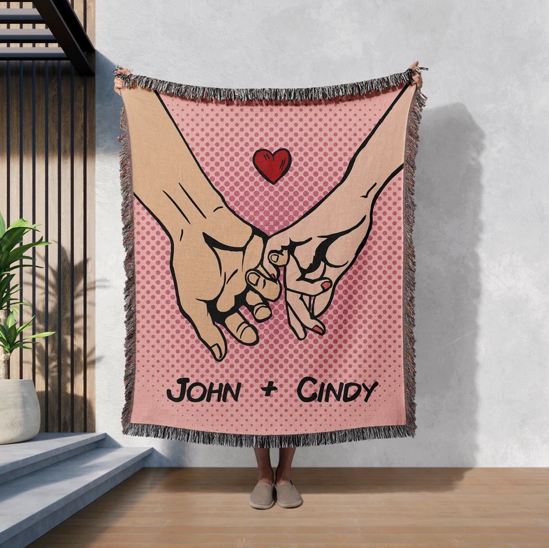 A pink blanket with a comic-style drawing of two hands clutching pinkies with a heart above them