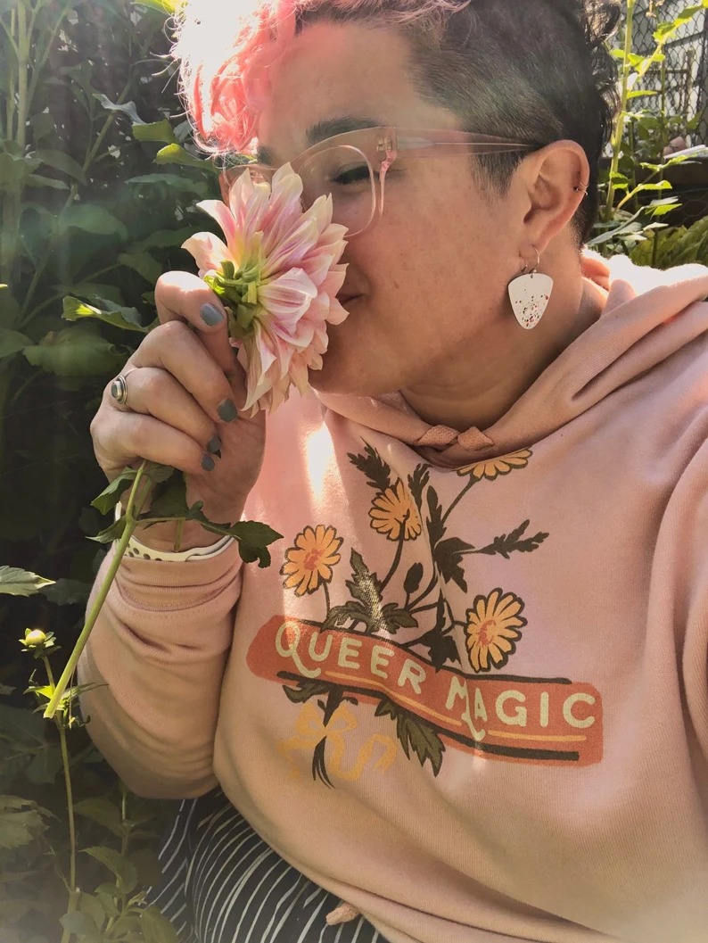 a photo of someone wearing a Queer Magic hoodie with illustrations of flowers smelling a flower