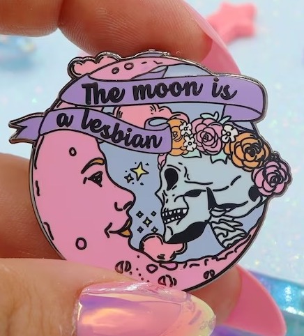 a pin with an illustration of a crescent moon and a skull wearing a flower crown facing each other with the text "the moon is a lesbian"
