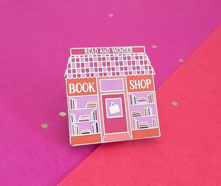 enamel pin in the shape of a bookshop on a pink background. 