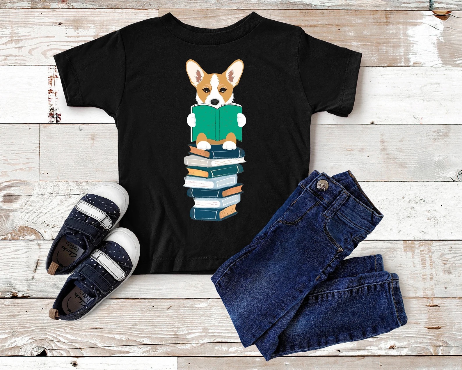 a photo of a black t-shirt featuring an illustration of a red and white Pembroke Welsh Corgi sitting on a stack of books. She is holding a book in her paws and is reading it.