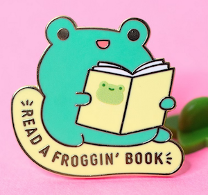 an enamel pin of an illustrated frog reading a book that says "read a froggin' book"