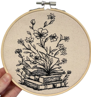 an embroidery hoop with a beige canvas and black thread to outline a stack of books and flowers