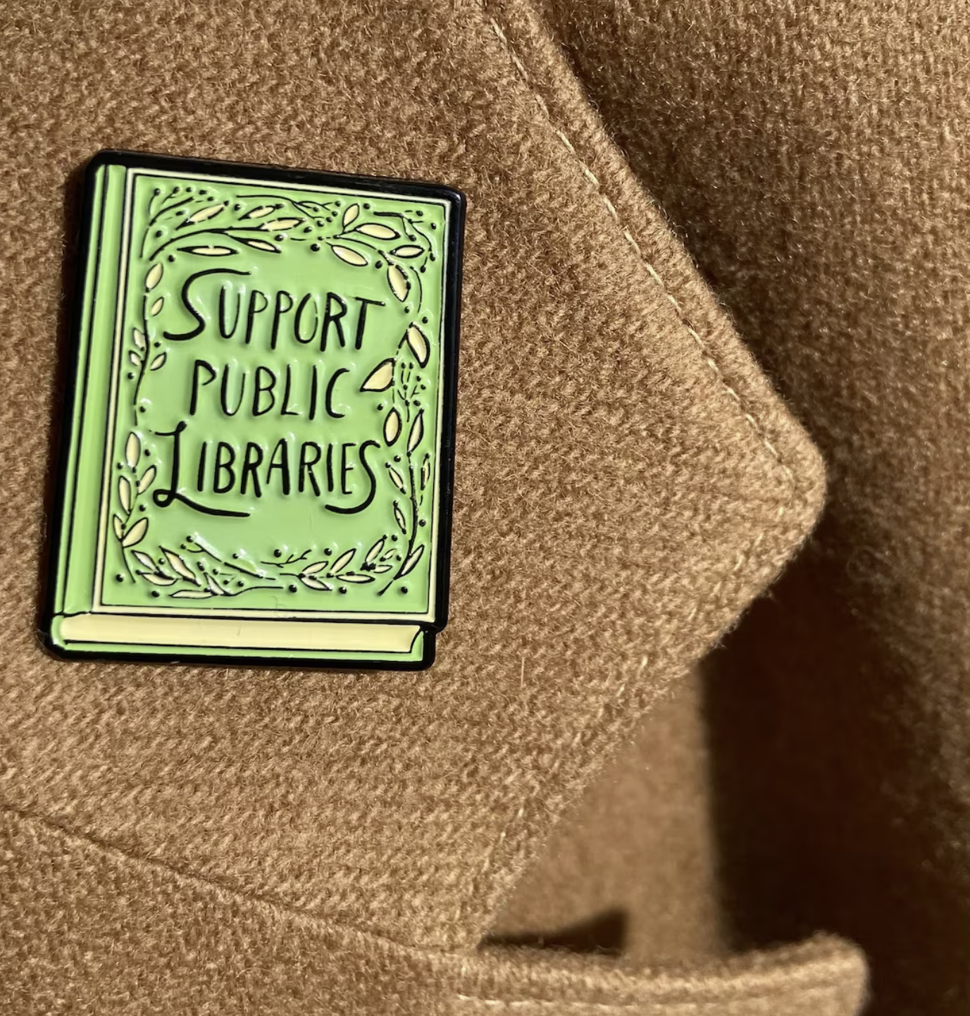 Lime Green enamel pin in the shape of a book with decorative leafy border around the edges and the text "support public libraries" is swirly font