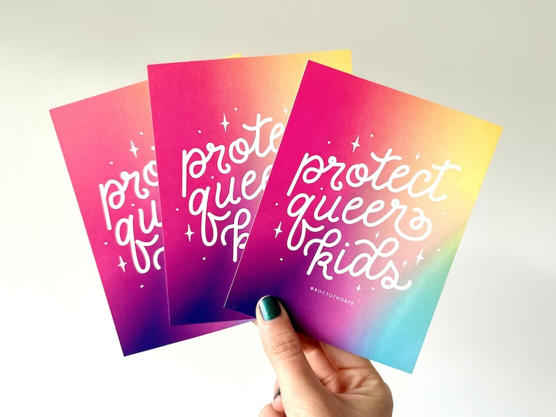 a photo of a hand holding up a set of colorful "protect queer kids" postcards