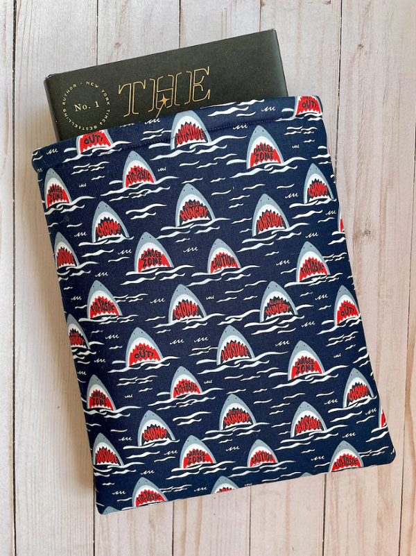 scary shark book sleve by mapleleafstitching a padded fabric book sleeve made with a patter of little shark heads, mouths open, and in the mouths it says things like chomp or dangerous waters