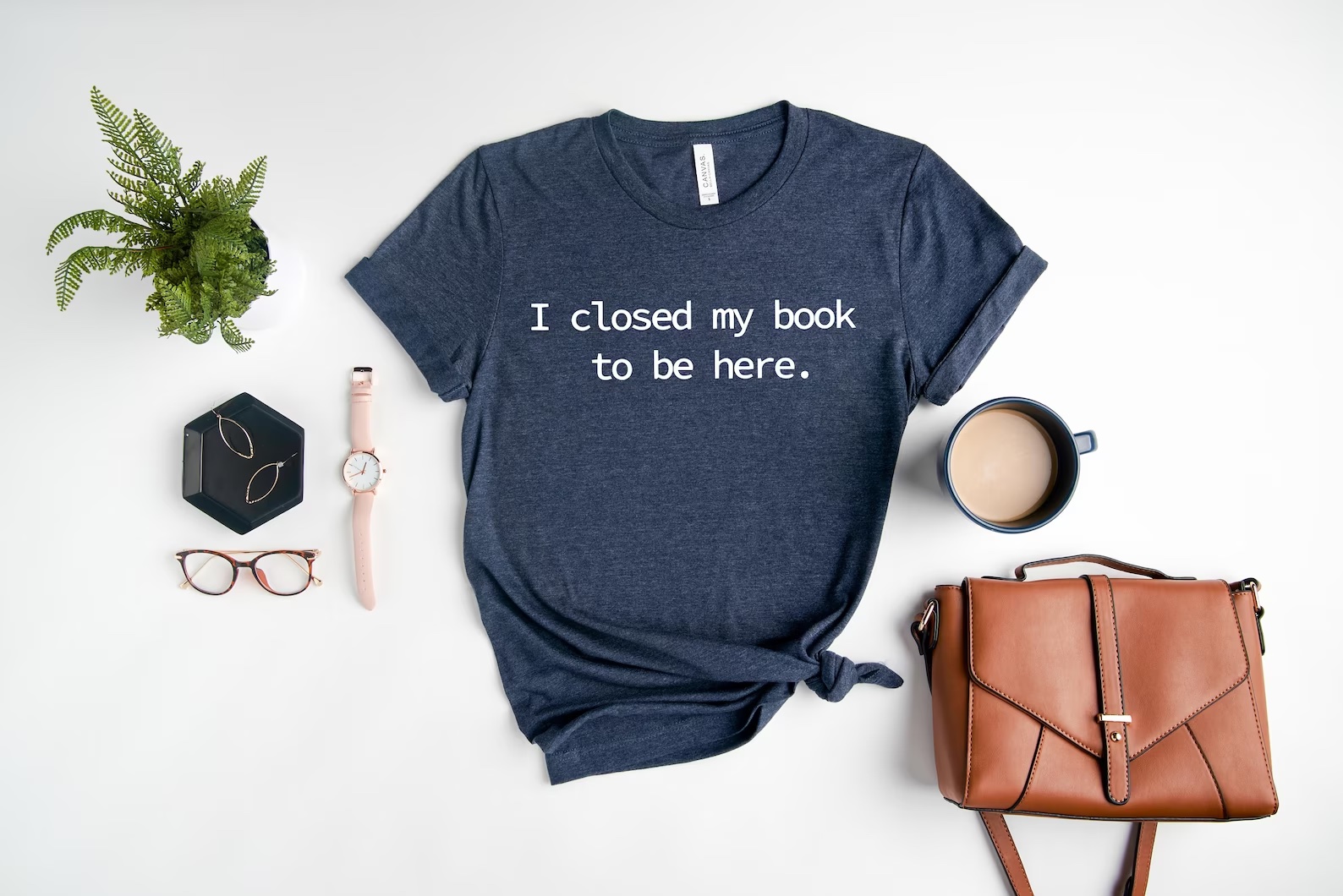 a photo of a blue t-shirt that says, "I closed my book to be here." The shirt is sitting next to a brown back and a plant.