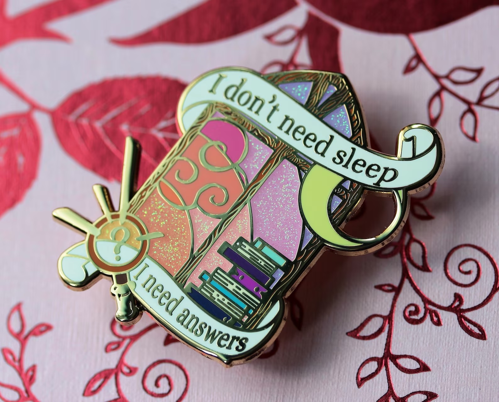 a photo of an enamel pin that features a multi-colored window near a stack of books. Text over the illustration read "I don't need sleep . . .  I need answers"
