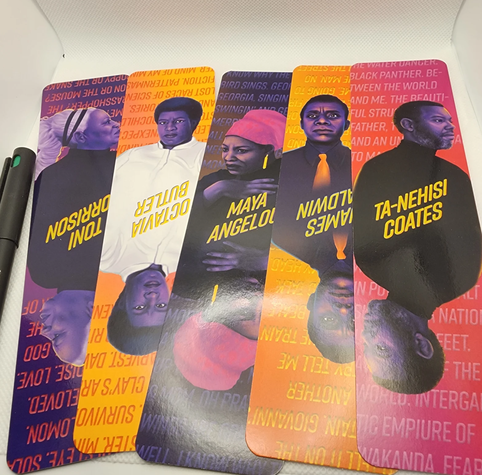 A colorful array of bookmarks in orange, purple, and bright pink featuring mirrored images of famous black authors including Octavia Butler, Toni Morrison, and James Baldwin. 
