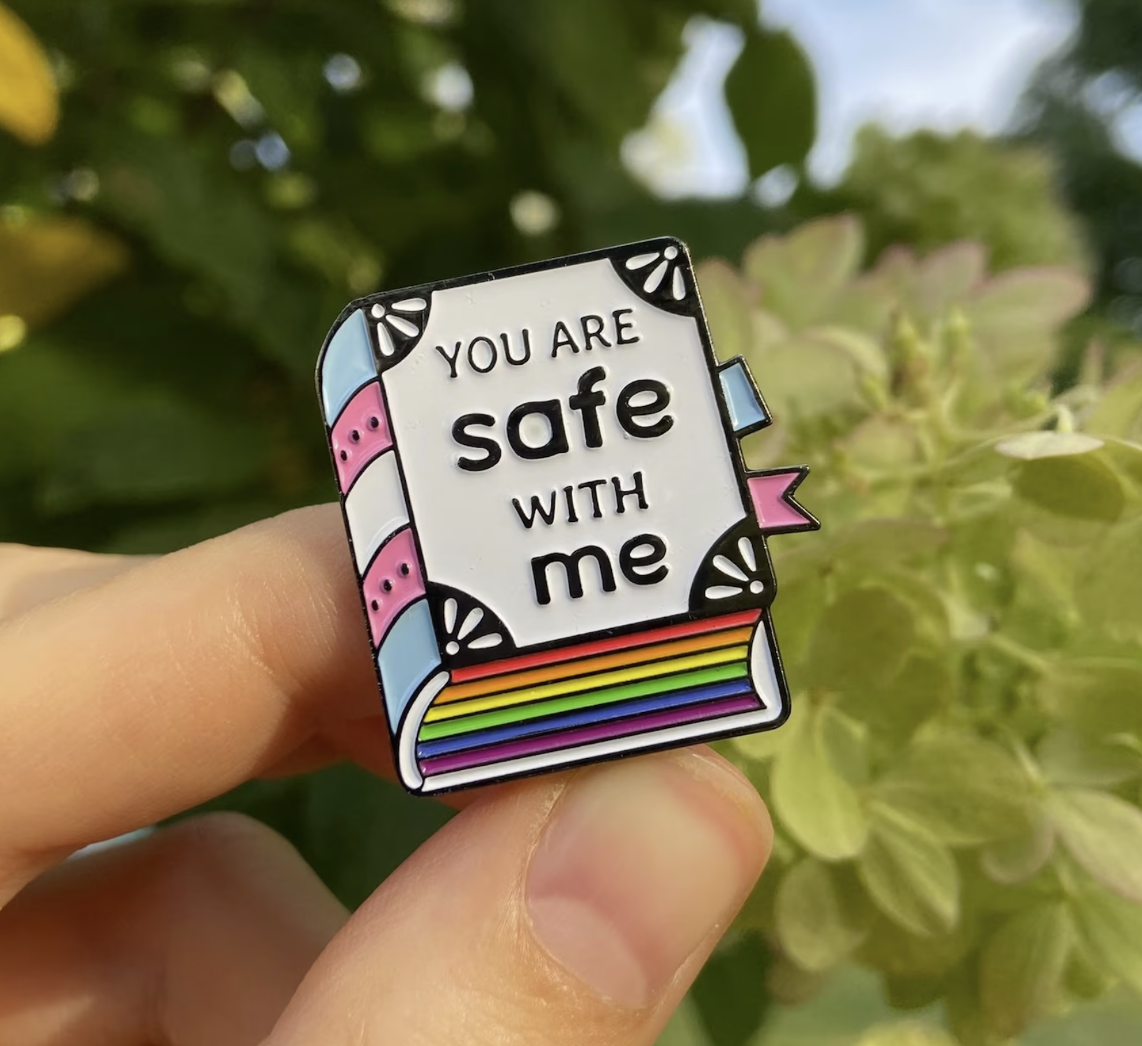 White hardcover book enamel pin with rainbow sprayed edges and the colors of the trans flag on the book spine. The words "You are safe with me" are written on the front cover. 