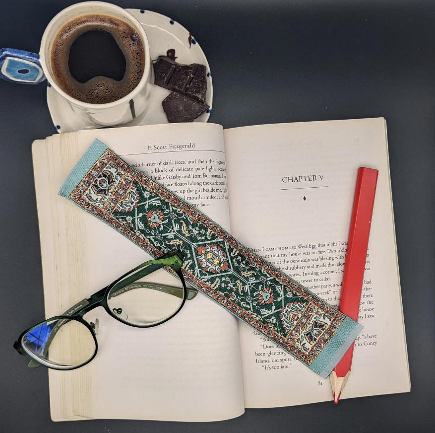 Tapestry bookmark in Turkish style in shades of green, blue, and read laid across an open book beside a cup of coffee. 