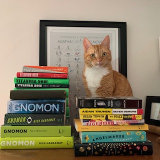 An orange cat sitting on top of a card catalog next to stacks of books by Nick Harkaway; photo by Liberty Hardy