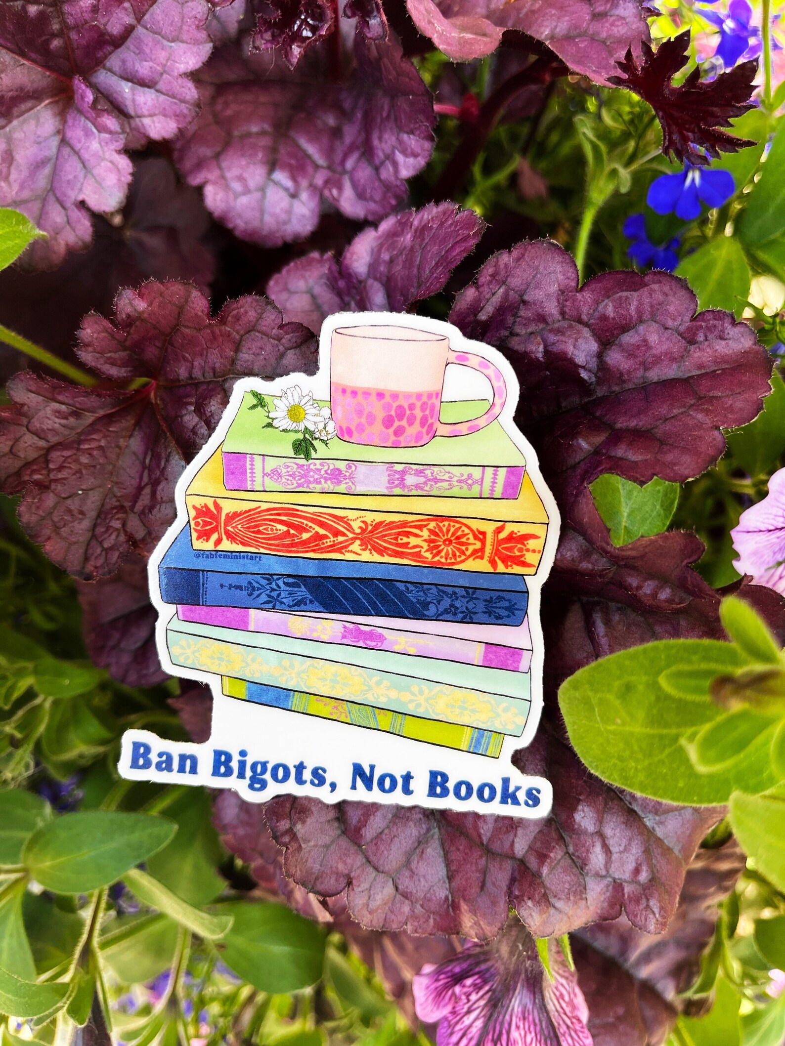 a photo of a sticker featuring an illustration of a colorful stack of books.  a teacup sits on the top of the stack. Words beneath the stack read, Ban bigots, not books