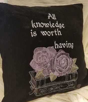 a photo of a black embroidered throw pillow with the text "All knowledge is worth having" with an image of a rose and a stack of books