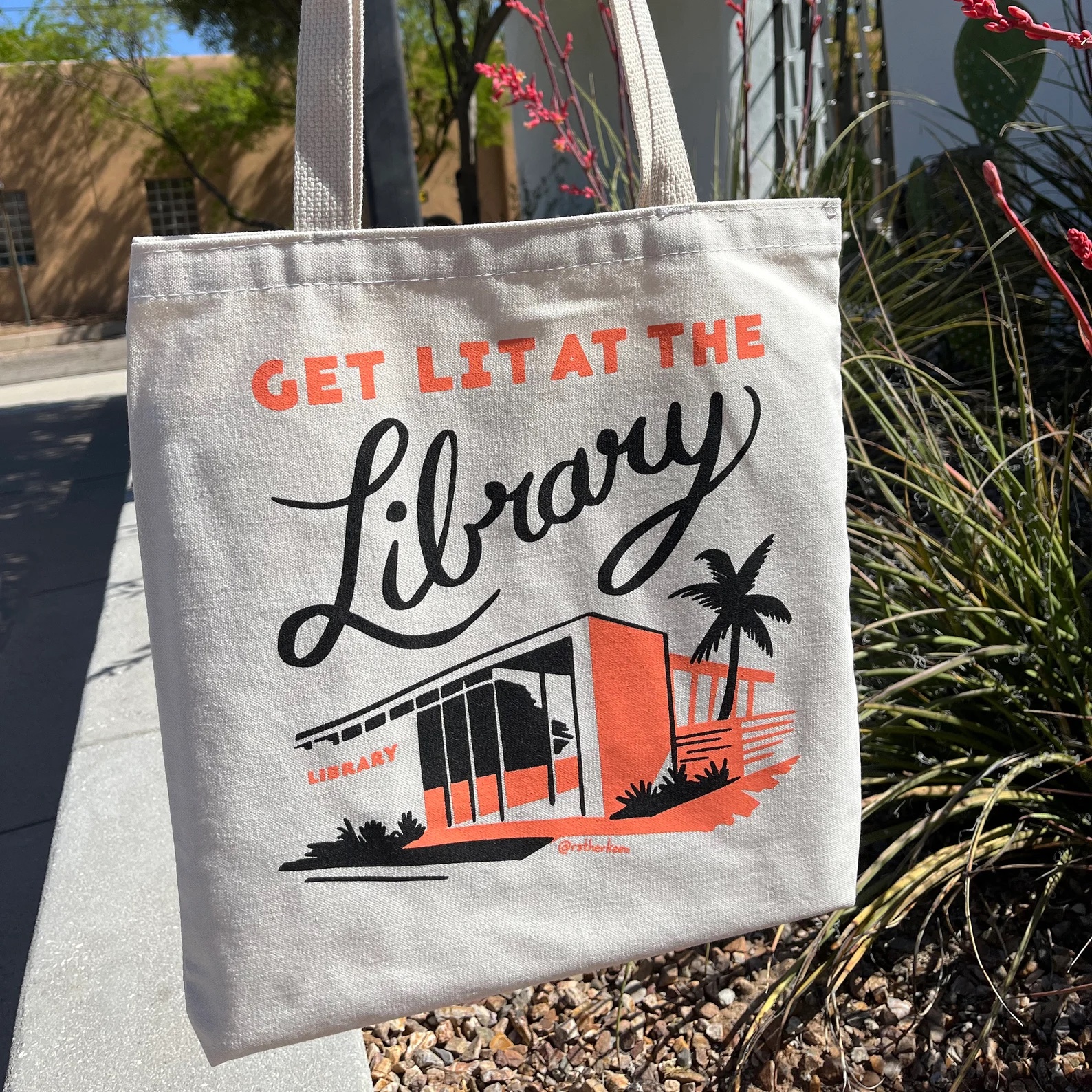 a photo of a canvas tote bag with an orange and black illustration of a library. The tote says, "Get Lit At The Library"