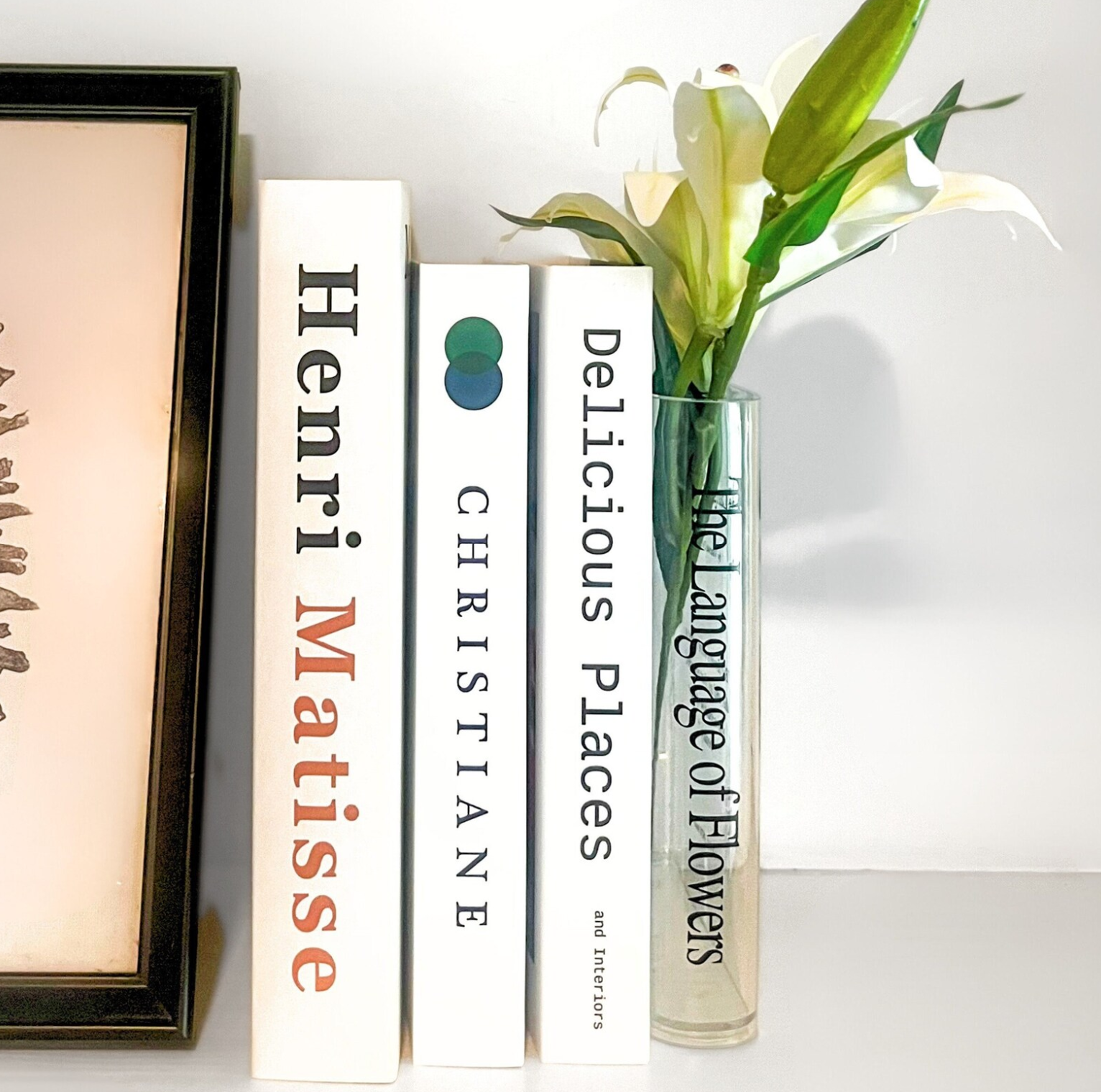 Picture of three white books lined up next to a clear acrylic vase in the shape of a book with green flowers sticking out of it.