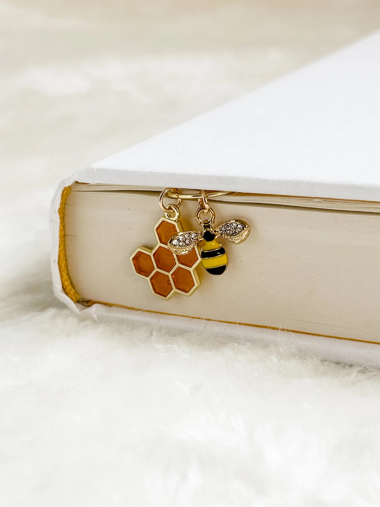 a photo of a bee bookmark. The book clips to a page and two charms hang over the book. One charm is a bee, and the other is honeycomb.