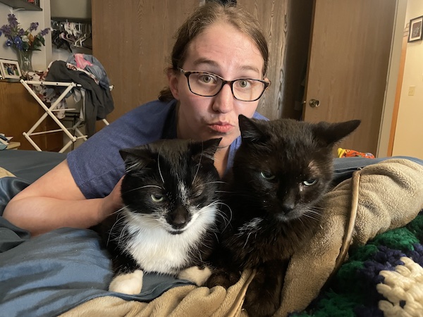 a brown haired woman making faces behind two cats sitting next to each other
