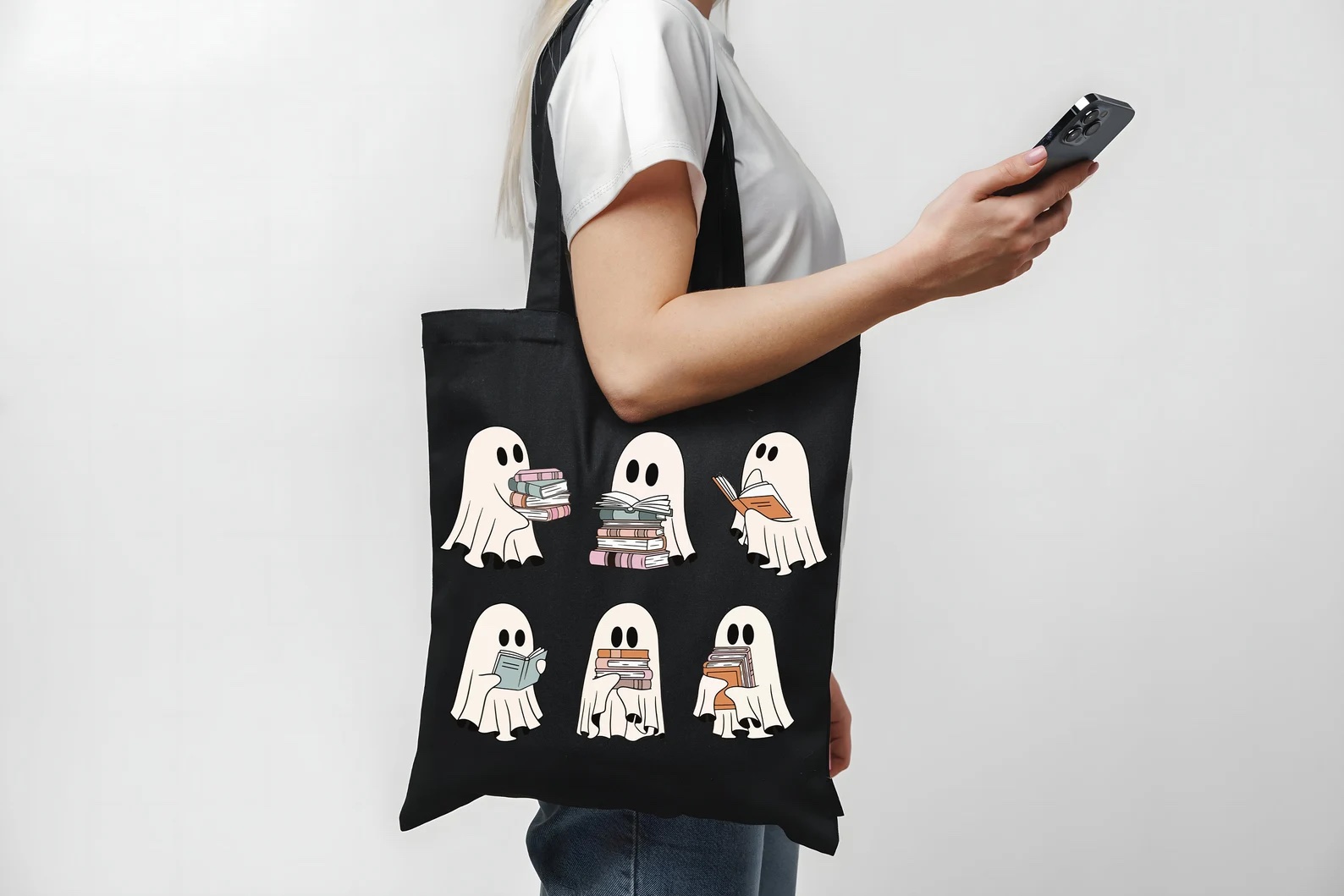 a photo of a white woman with a navy tote bag on her shoulder. The tote bag features six ghosts in various poses reading and carrying books.