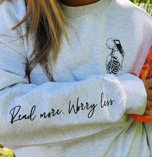 sweatshirt with skeleton holding book illustration on chest and down sleeve printed text "read more worry less"