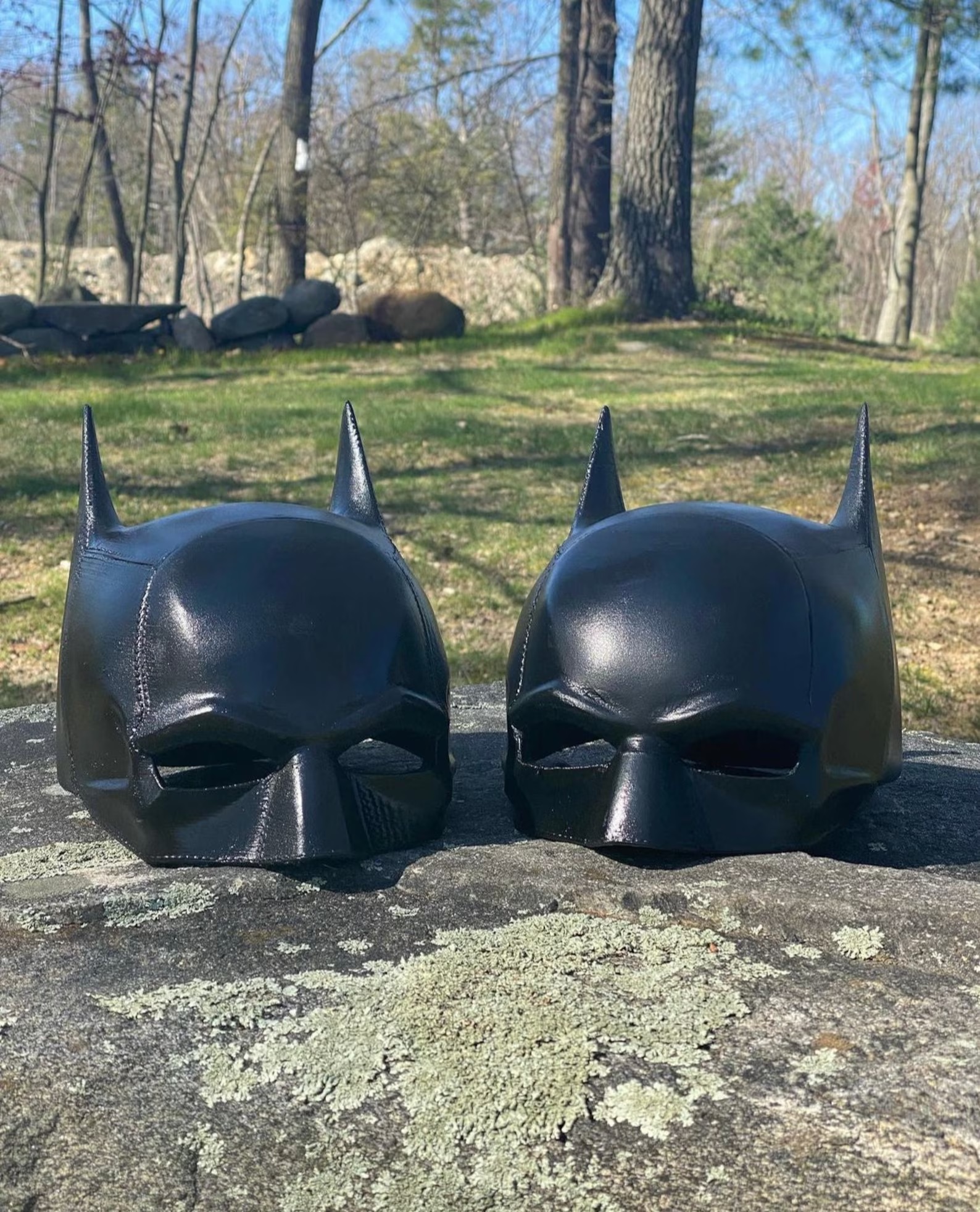 Two Batman cowls sitting side by side on a rock