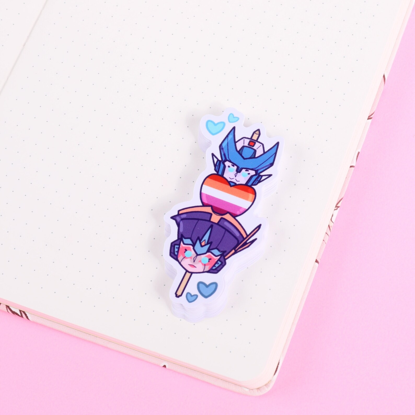 A sticker with the Transformers characters Chromia and Windblade stacked atop each other with a lesbian pride heart in between
